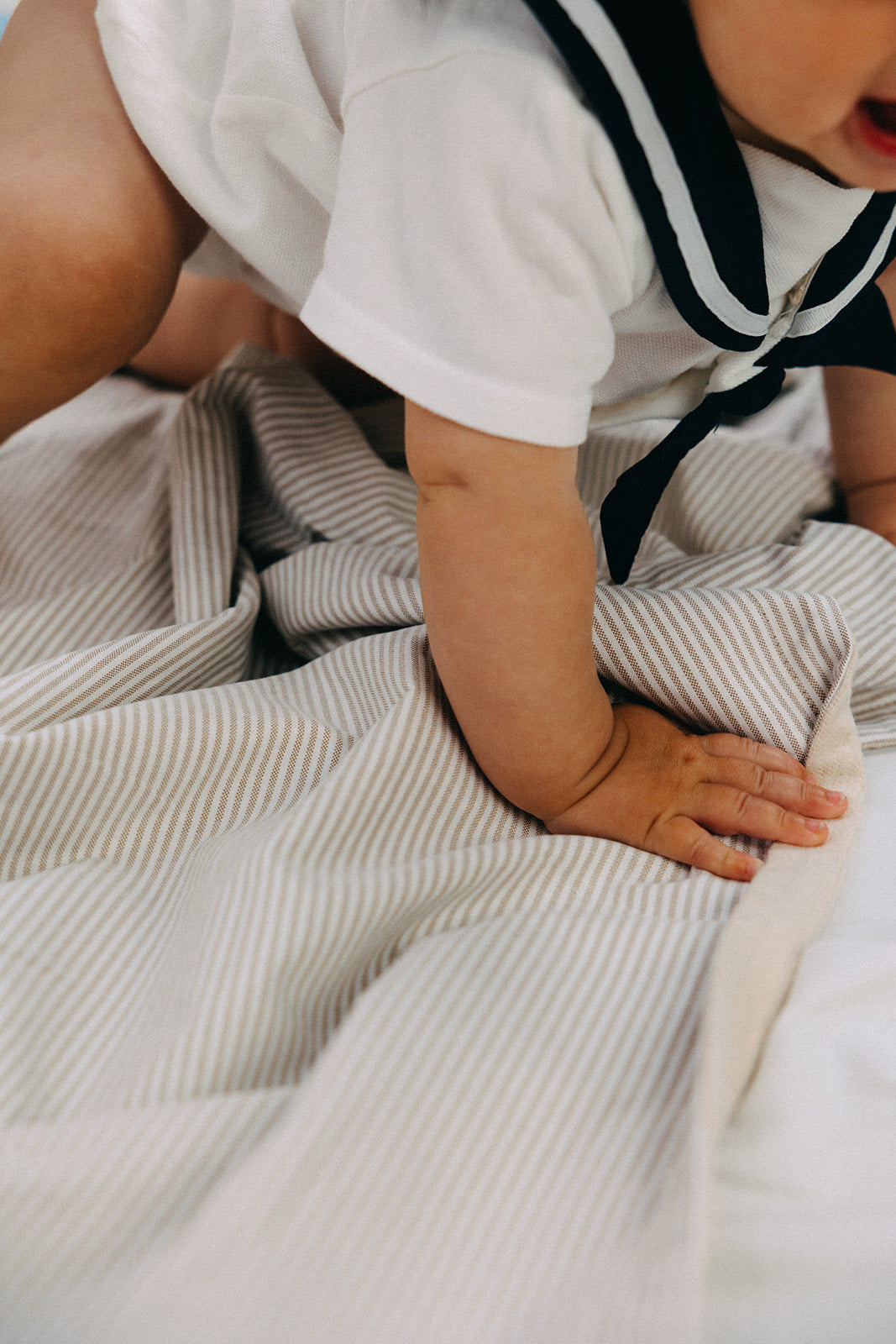 A close-up lifestyle shot of a baby crawling on a cotton white and beige Oxford striped blanket, wearing a white short-sleeve top with a navy and white sailor collar.