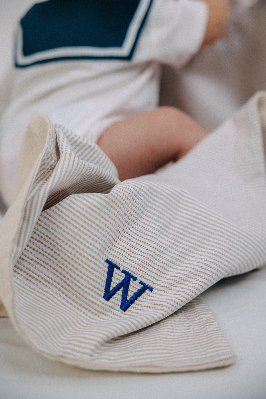 A close-up of a baby's legs and torso wrapped in a cream and white Oxford striped blanket with a blue embroidered letter 'W', wearing a white and navy sailor-themed bodysuit