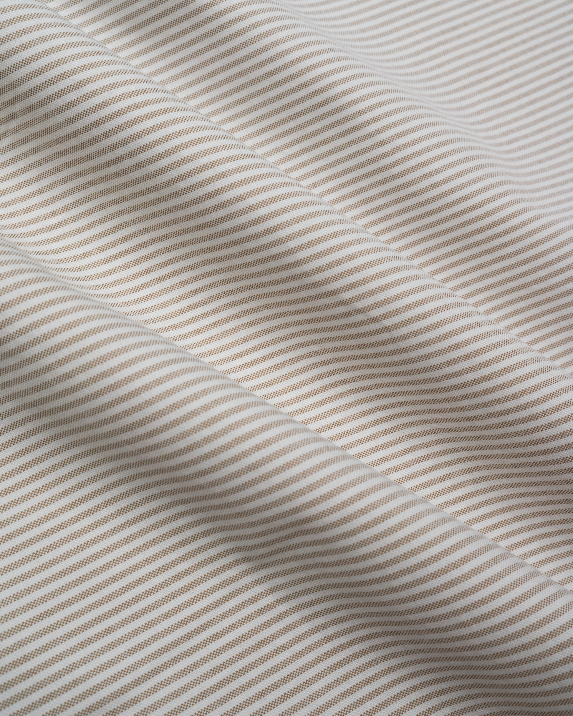 A close-up of a woven fabric with beige and white Oxford stripes, showing soft folds and texture