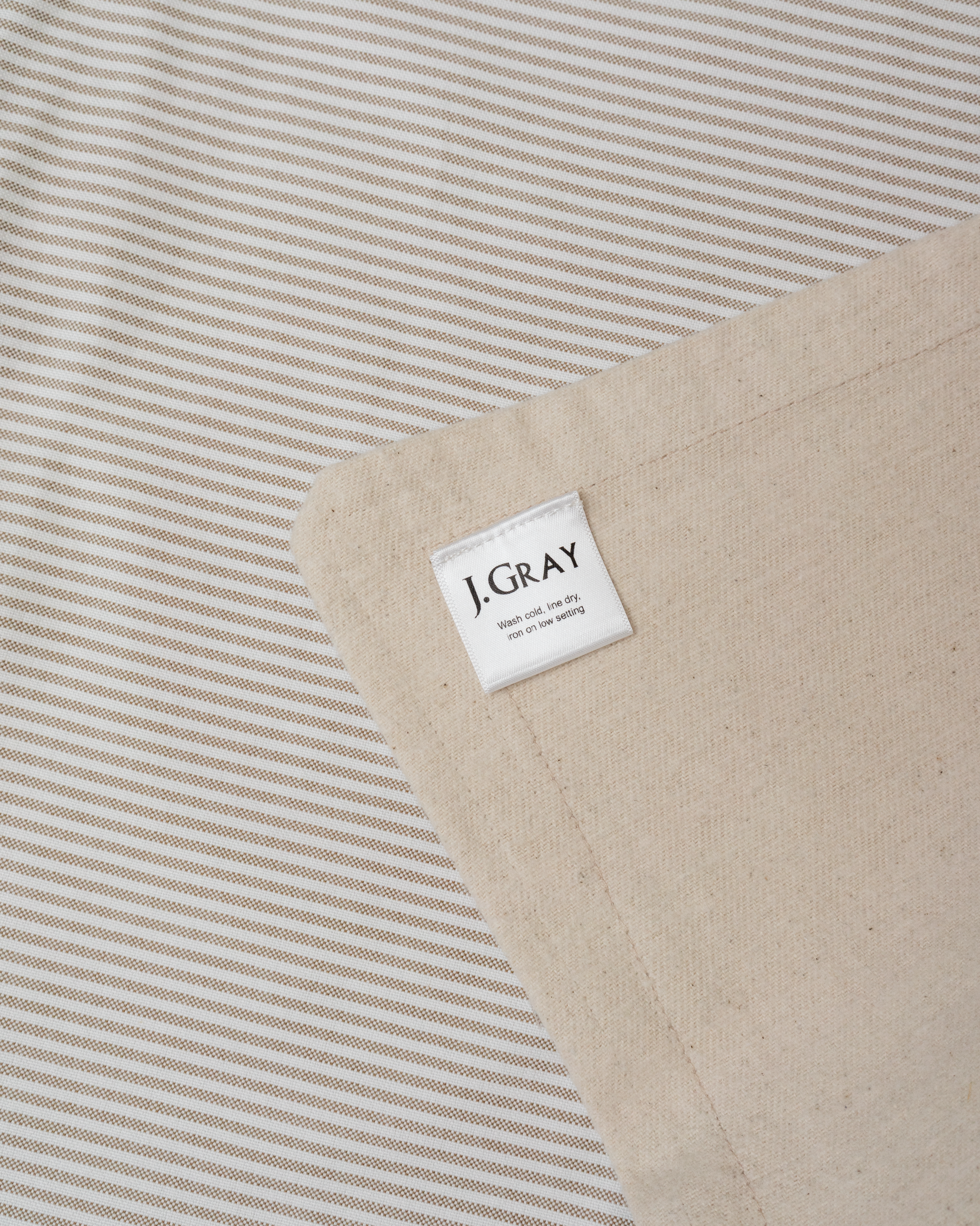 Close-up of a J. Gray baby blanket corner showing a cream woven label on a beige fabric with Oxford stripes
