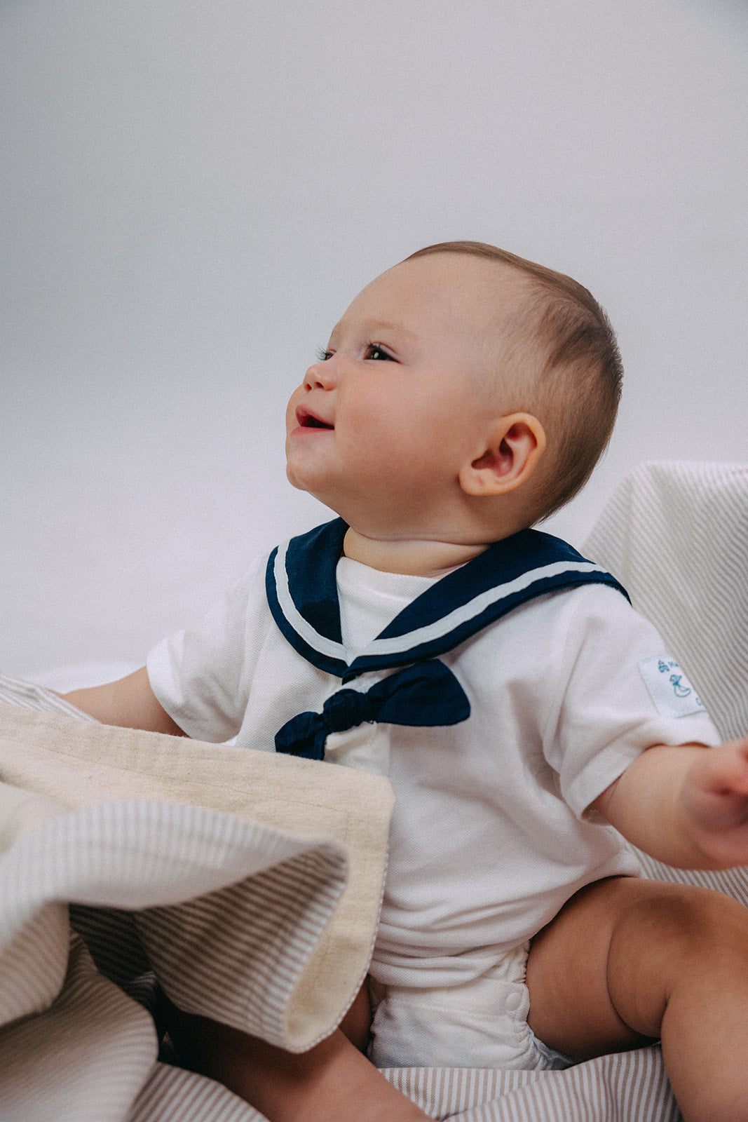 A baby wearing a white short-sleeve bodysuit with a navy blue sailor collar, sitting on a beige and white Oxford striped blanket against a light gray background