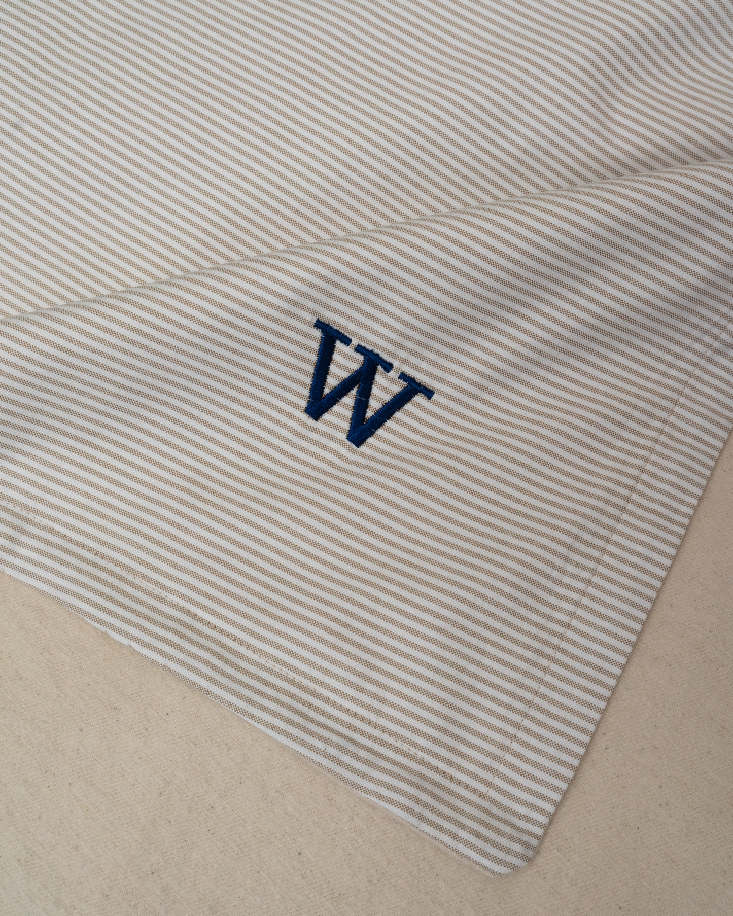 Close-up of a cotton baby blanket, featuring thin beige and white Oxford stripes with a navy blue embroidered letter 'W' in the corner
