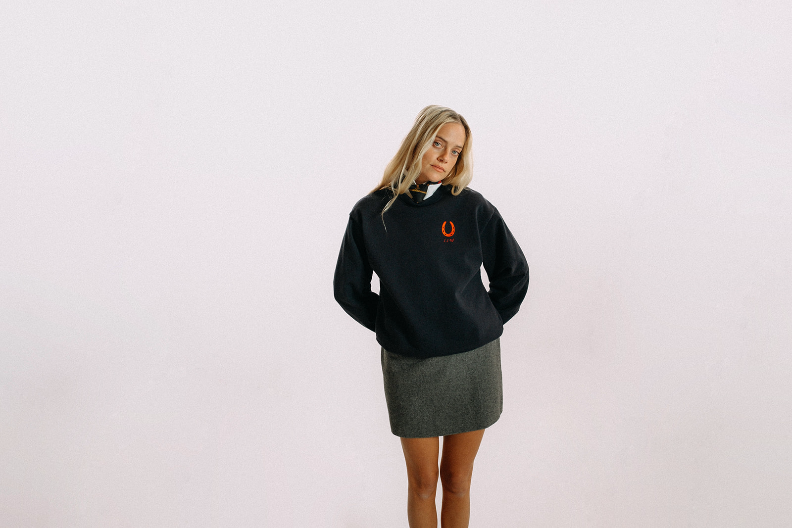 Woman wearing navy blue sweater with red horseshoe at left chest, white collared shirt, tie and grey skirt